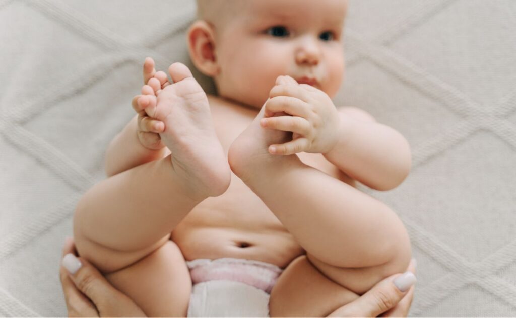 Baby wearing only a nappy and holding their feet while mother's hands hold baby's legs