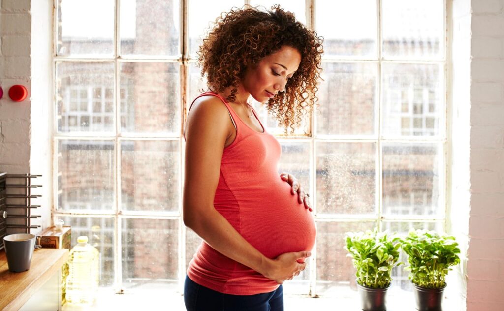 A pregnant woman holds her bump thoughtfully in her modern kitchen window on a sunny day