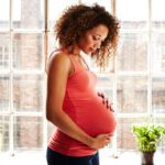 A pregnant woman holds her bump thoughtfully in her modern kitchen window on a sunny day