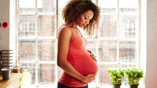 A pregnant woman holds her bump thoughtfully in her modern kitchen window on a sunny day