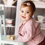 Old fashioned baby names: A beautiful baby girl, 12 months old, smiles at the camera as she stands against a child-sized wooden chair. Shabby chic cupboards of vintage tea cups are in the background.