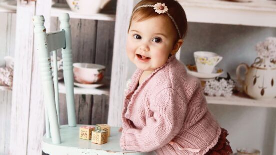 Old fashioned baby names: A beautiful baby girl, 12 months old, smiles at the camera as she stands against a child-sized wooden chair. Shabby chic cupboards of vintage tea cups are in the background.