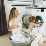 Beautiful mother and her daughter enjoying and laughing doing the laundry in the kitchen of modern apartment where a washing machine can be seen on. They doing household chores together, they smiling and toddler girl helping her parent.