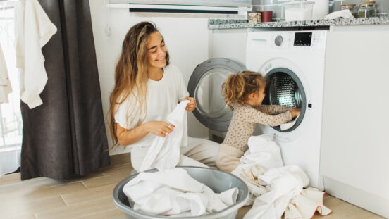 Beautiful mother and her daughter enjoying and laughing doing the laundry in the kitchen of modern apartment where a washing machine can be seen on. They doing household chores together, they smiling and toddler girl helping her parent.