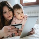 young mother with a baby girl lying on the bed and online with the tablet and holding her credit card in hand