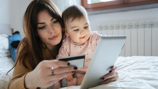 young mother with a baby girl lying on the bed and online with the tablet and holding her credit card in hand