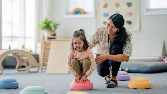 A female Physiotherapist works with a young girl on her balancing during a therapy session. She is supporting and aiding the young girl as they work together.